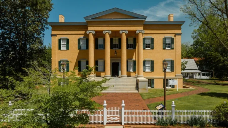 A large, historic, two-story yellow house with six grand columns supporting the entrance, surrounded by a white fence and lush greenery. The house features multiple windows with shutters and chimneys on either side. A brick pathway leads to the main entrance.