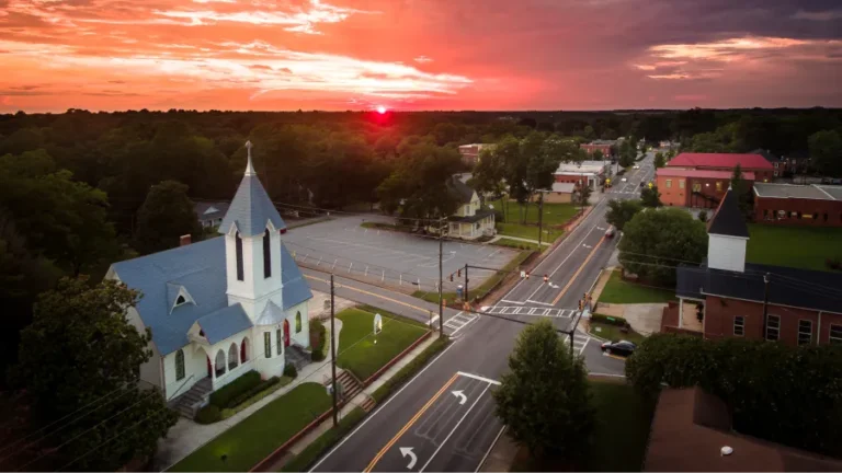 Aerial view of a quaint small town at sunset, featuring a charming white church with a blue roof in the foreground. The street is lined with trees, houses, and small buildings, bathed in the warm, glowing light of the setting sun.