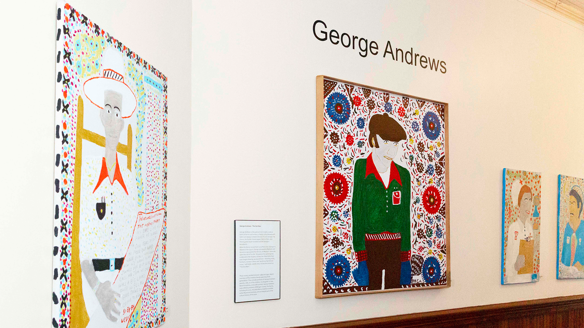 An art gallery wall displays several colorful paintings with different patterns by George Andrews. Each painting features stylized human figures with detailed outfits and backgrounds inspired by Great Stories from small towns. A plaque with text is placed between the artworks.