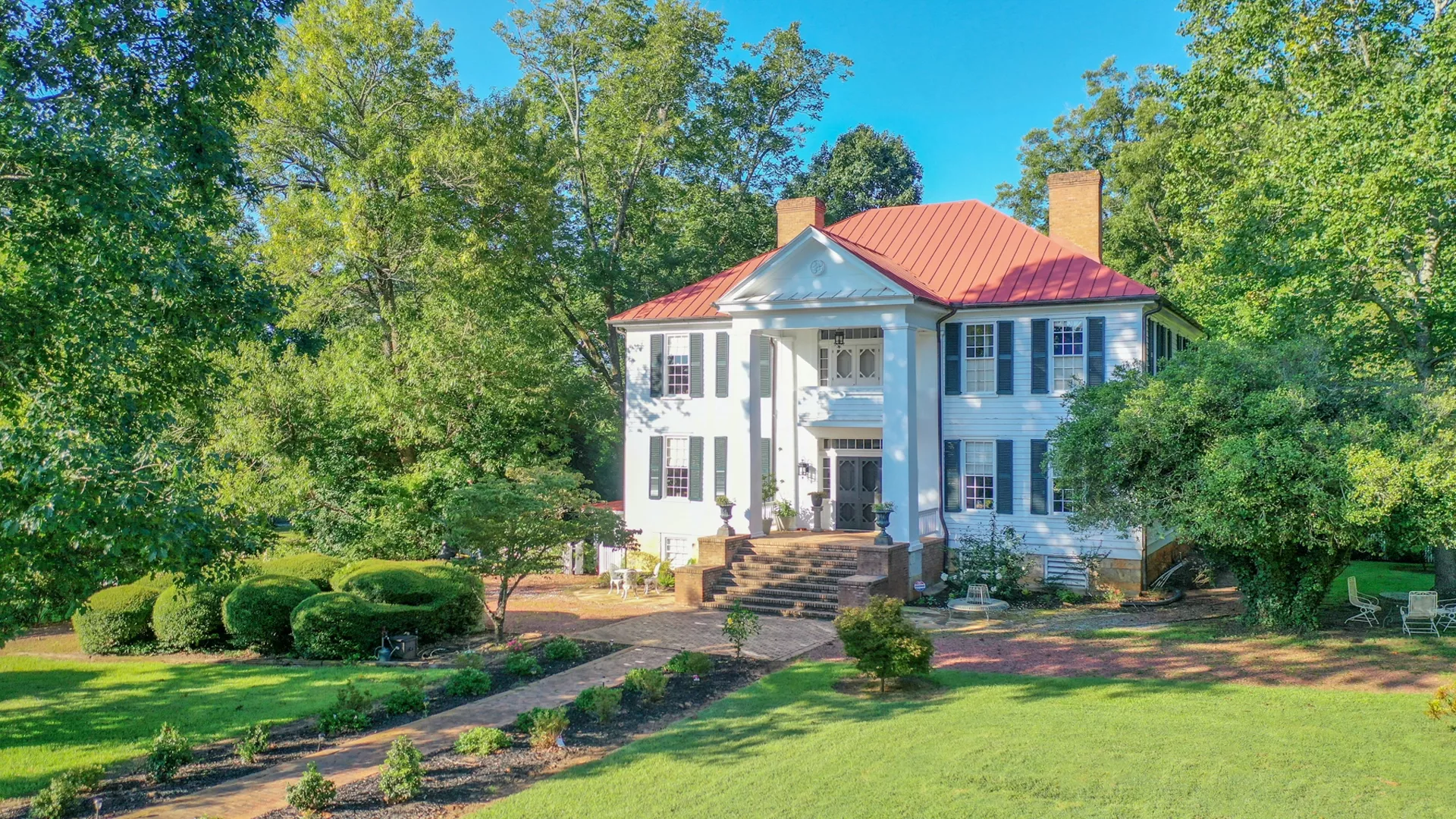 A large, historic accommodation in the form of a white two-story house with a red roof is surrounded by lush greenery. The house features tall columns, a grand entrance with steps leading up to it, and multiple windows. The front yard includes manicured lawns, bushes, and trees.