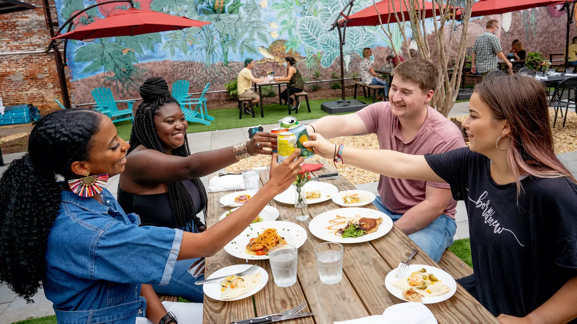 A group of four people, two women and two men, are sitting around a wooden table outdoors, enjoying a meal. They are raising their drinks in a toast and smiling. Plates of food and glasses of water are on the table, with a colorful mural in the background—an ideal scene for any dining guide on where to dine.